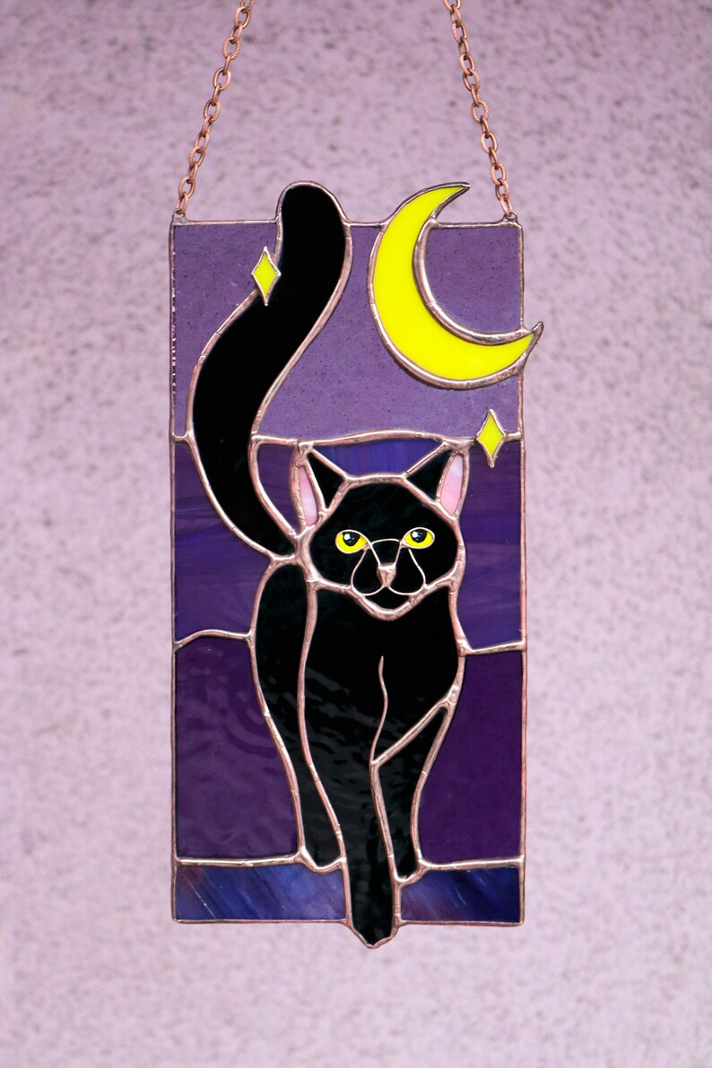 May include: A stained glass window hanging with a black cat silhouette walking in front of a purple night sky with a yellow crescent moon and stars.