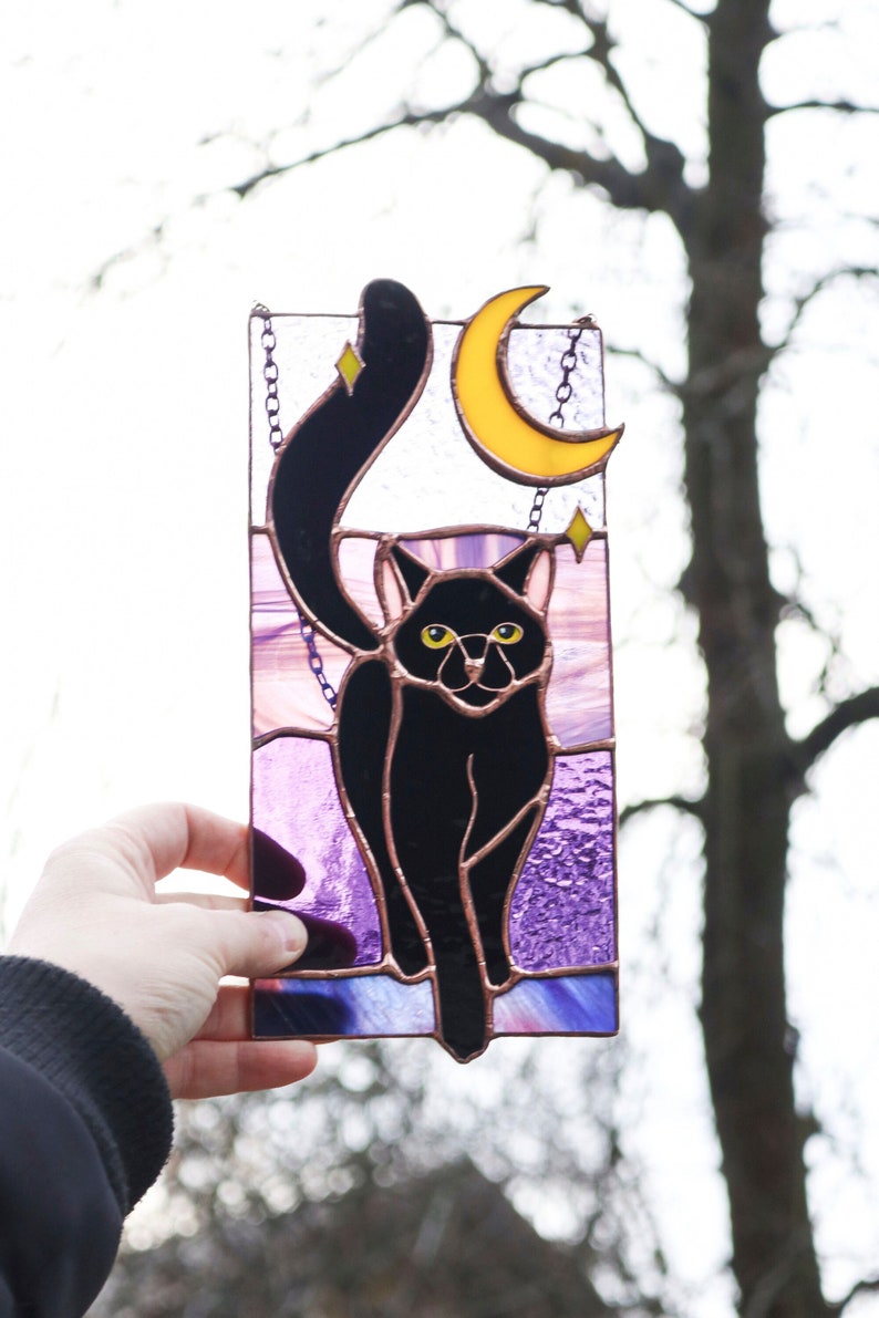 May include: A stained glass window hanging features a black cat with yellow eyes standing in front of a crescent moon. The background is purple and pink.