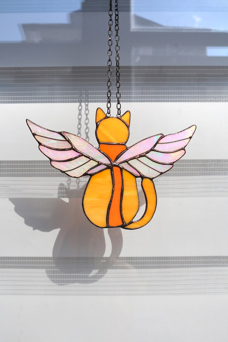May include: A stained glass cat with iridescent wings hanging from a chain. The cat is orange and the wings are pink and white.