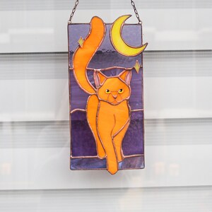 May include: A stained glass window hanging with a chain. The window features an orange cat with a crescent moon and stars in the background.