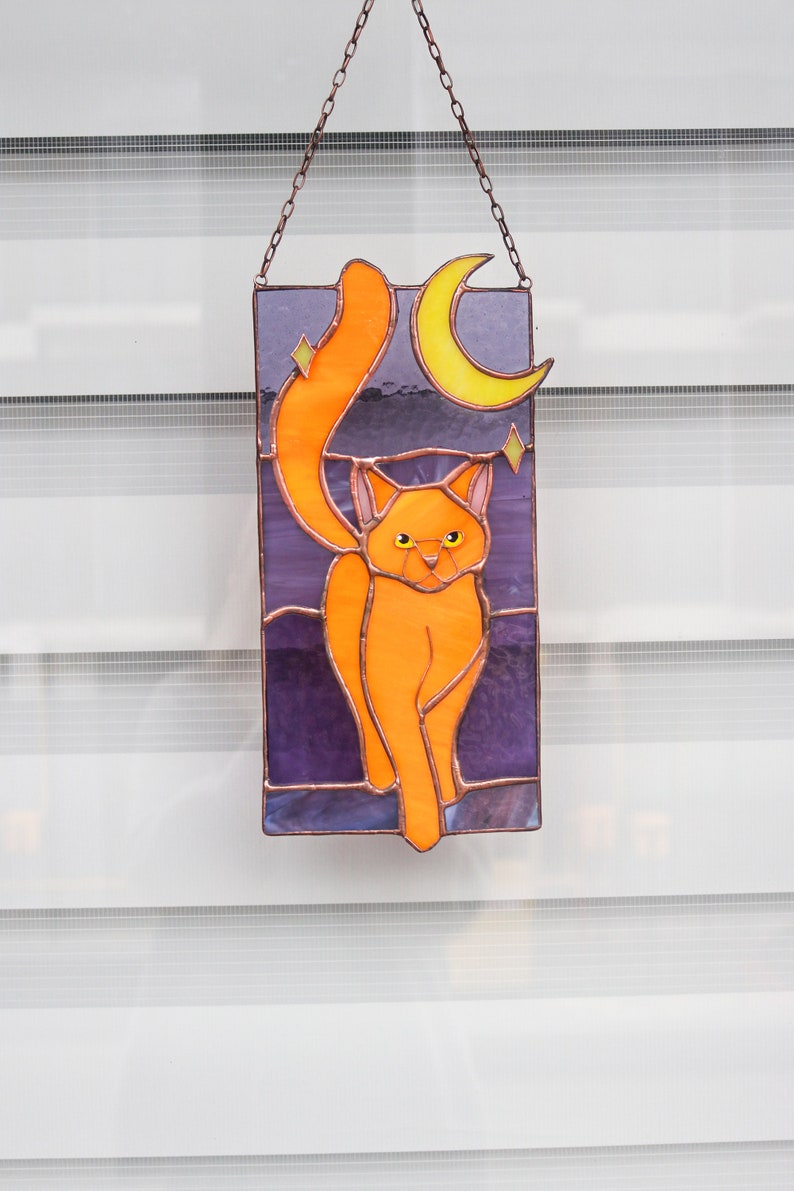 May include: A stained glass window hanging with a chain. The window features an orange cat with a crescent moon and stars in the background.