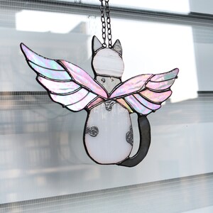 May include: A stained glass cat with iridescent wings hanging from a black chain. The cat is white with black spots and has a black tail. The wings are pink and purple.