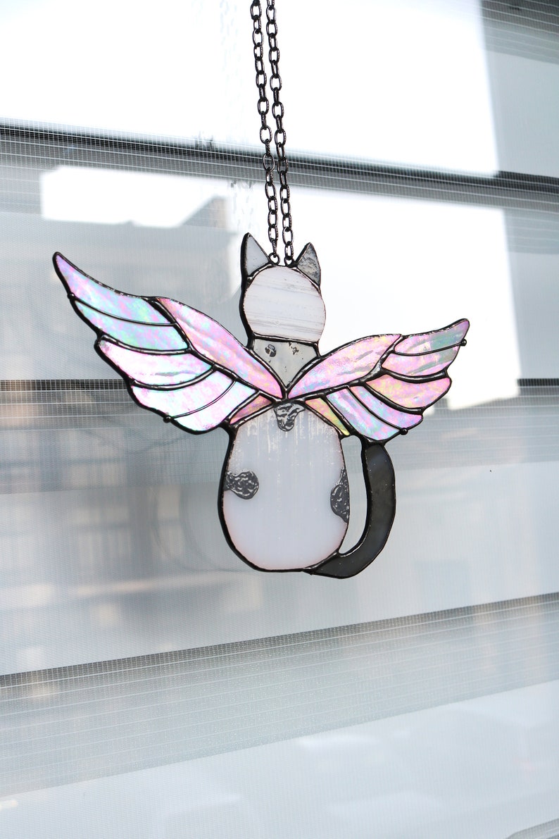 May include: A stained glass cat with iridescent wings hanging from a black chain. The cat is white with black spots and has a black tail. The wings are pink and purple.