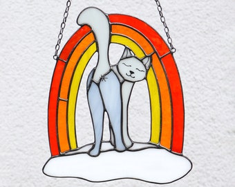 May include: A stained glass cat hanging from a chain, standing on a cloud in front of a rainbow. The cat is white with a grey body and a rainbow with red, orange, yellow, and green stripes.
