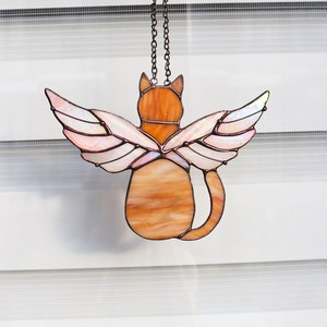 May include: A stained glass cat with wings hanging from a chain. The cat is orange and brown with pink and white wings.