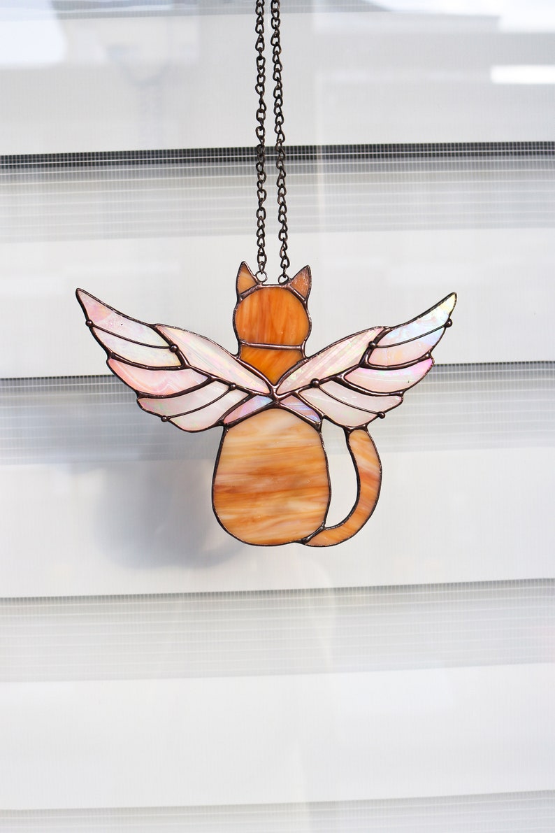 May include: A stained glass cat with wings hanging from a chain. The cat is orange and brown with pink and white wings.
