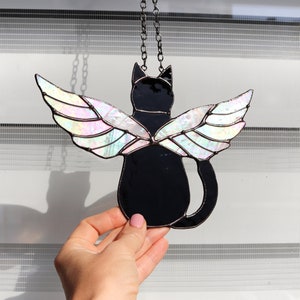 May include: A stained glass window hanging with a black cat with iridescent wings. The cat is facing away from the viewer and the wings are spread out behind it.