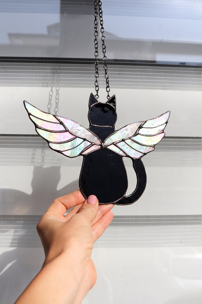 May include: A stained glass window hanging with a black cat with iridescent wings. The cat is facing away from the viewer and the wings are spread out behind it.