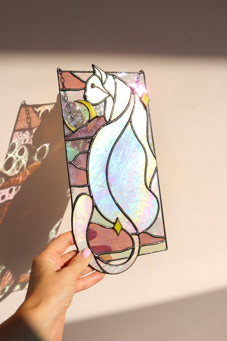 May include: A stained glass window hanging depicts a white cat with iridescent glass, sitting in front of a pink and yellow moon. The cat's tail curls around the moon.