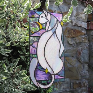 May include: A stained glass window hanging from a chain, depicting a white cat with iridescent glass, sitting in front of a crescent moon and stars. The background is a purple, blue, and green gradient.