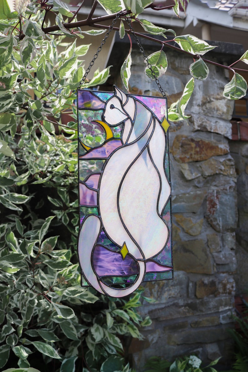 May include: A stained glass window hanging from a chain, depicting a white cat with iridescent glass, sitting in front of a crescent moon and stars. The background is a purple, blue, and green gradient.