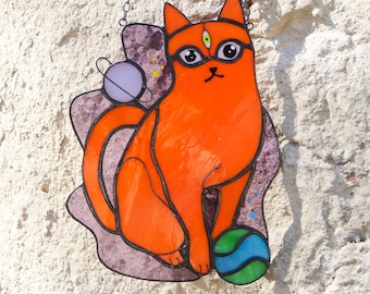 May include: Stained glass cat with an orange body, sitting on a purple base. The cat has a third eye and is holding a blue and green ball. A planet with rings is to the left. The cat is hanging from a chain.