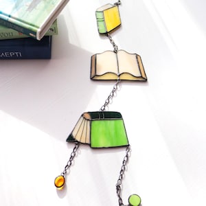 Peut inclure: Mobile en vitrail représentant des livres, avec trois formes de livres en jaune, crème et vert, reliées par des chaînes. Il comporte des accents de perles de verre orange et vertes. Fond blanc, avec des livres en arrière-plan.
