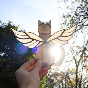 May include: A stained glass cat with wings, the cat is light brown and the wings are iridescent with a pink and white color scheme. The cat is being held up in front of a bright blue sky and trees.