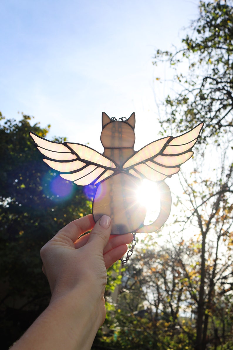 May include: A stained glass cat with wings, the cat is light brown and the wings are iridescent with a pink and white color scheme. The cat is being held up in front of a bright blue sky and trees.