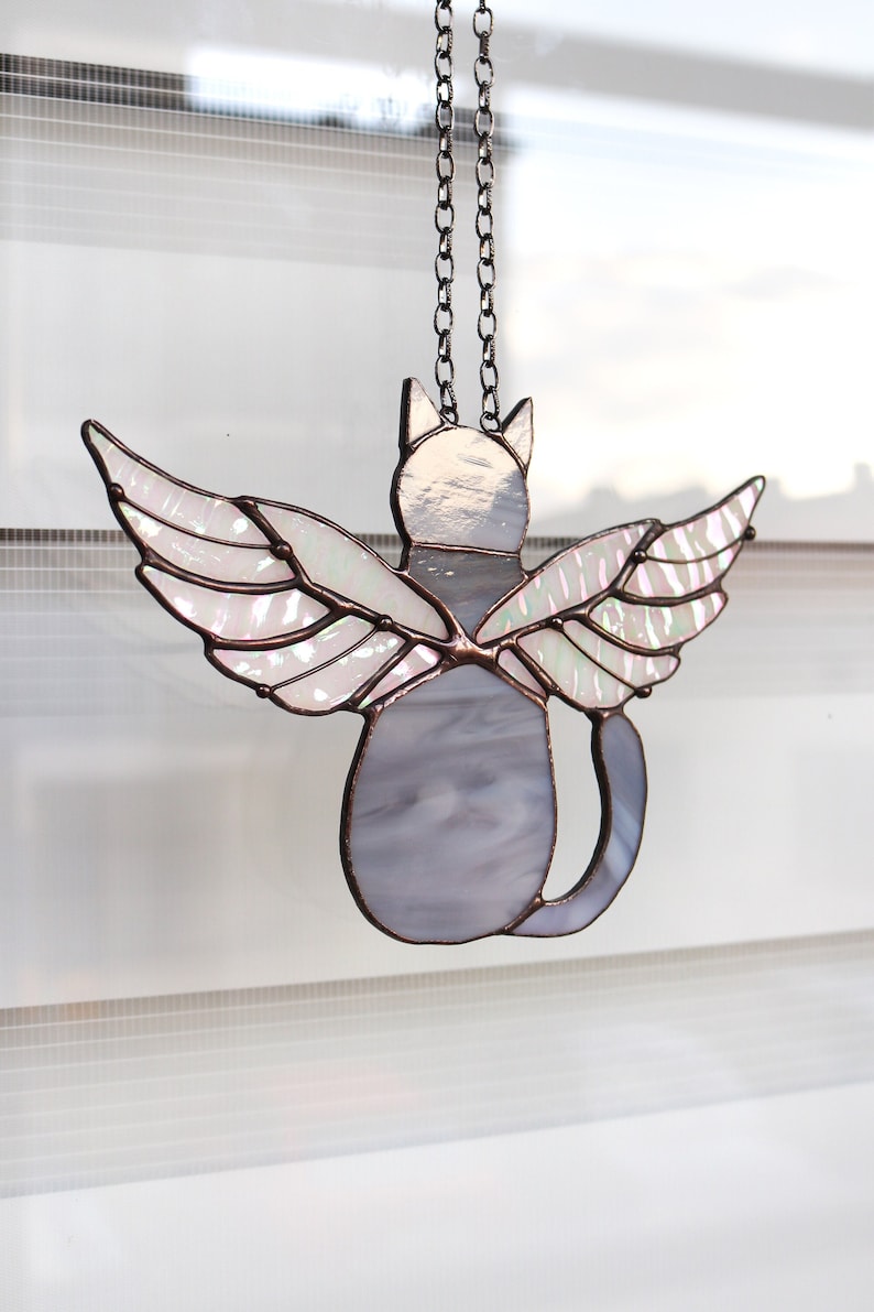 May include: A stained glass cat with iridescent wings hanging from a chain. The cat is gray and the wings are white with a pink iridescent sheen.