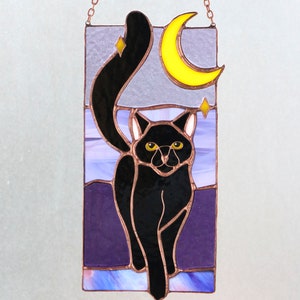 May include: A stained glass window hanging with a black cat walking towards the viewer. The cat has yellow eyes and is silhouetted against a purple and blue background with a yellow crescent moon and stars.
