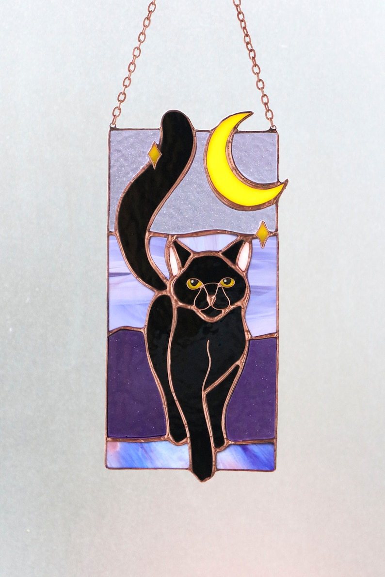 May include: A stained glass window hanging with a black cat walking towards the viewer. The cat has yellow eyes and is silhouetted against a purple and blue background with a yellow crescent moon and stars.