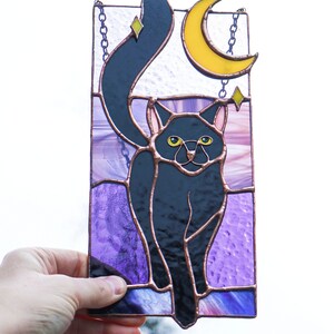 May include: A stained glass window hanging with a black cat silhouette with yellow eyes, a crescent moon, and stars on a purple background.