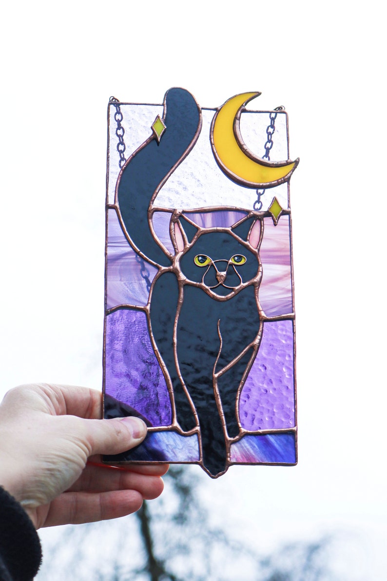 May include: A stained glass window hanging with a black cat silhouette with yellow eyes, a crescent moon, and stars on a purple background.