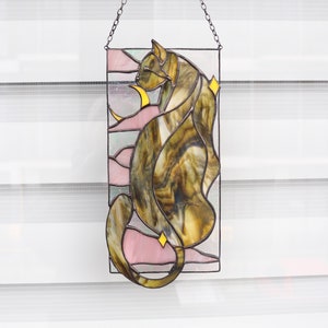May include: A stained glass window hanging from a chain. The window depicts a brown and yellow cat with pink and white accents.