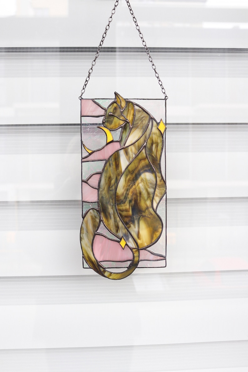 May include: A stained glass window hanging from a chain. The window depicts a brown and yellow cat with pink and white accents.