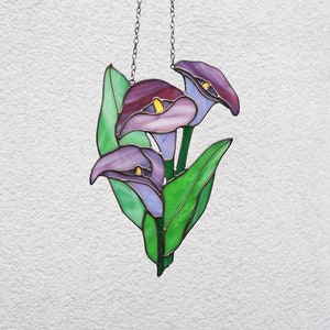 May include: A stained glass window hanging with a chain. The window features three purple calla lilies with green leaves.