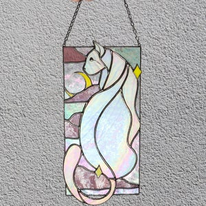 May include: A stained glass window hanging from a chain. The window features a white cat with a crescent moon and stars in the background. The cat is outlined in black and the background is a mix of pink, purple, and blue.