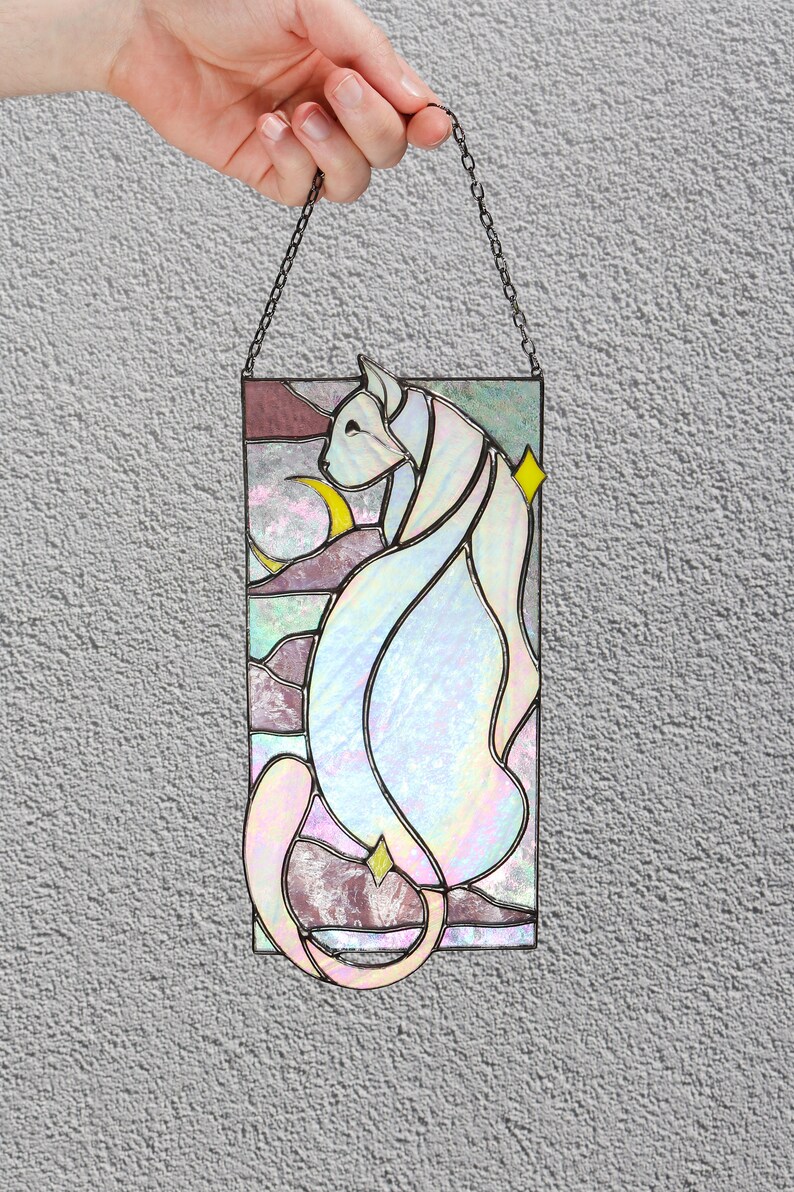 May include: A stained glass window hanging from a chain. The window features a white cat with a crescent moon and stars in the background. The cat is outlined in black and the background is a mix of pink, purple, and blue.