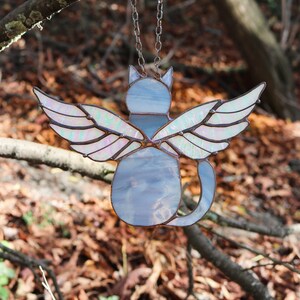 May include: A stained glass cat with iridescent wings hanging from a silver chain. The cat is light blue and the wings are iridescent with pink and green hues. The cat is facing away from the viewer.