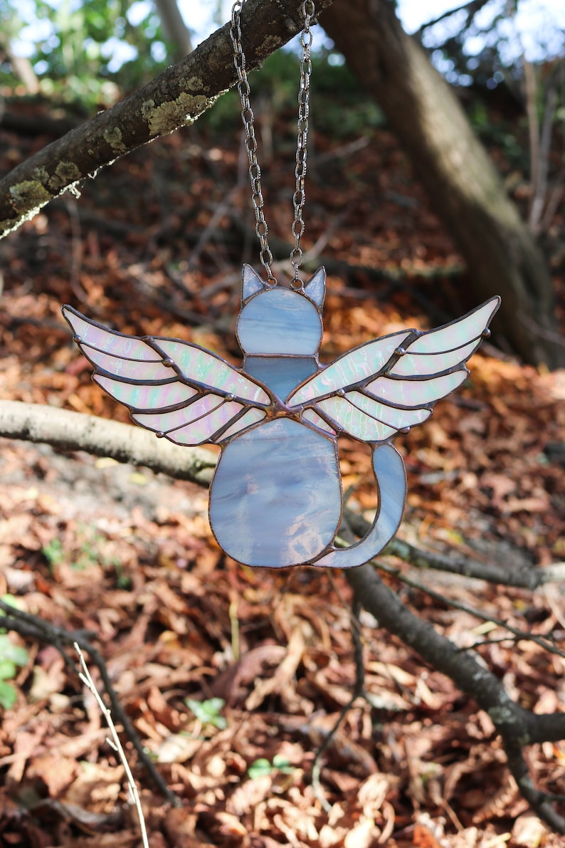 May include: A stained glass cat with iridescent wings hanging from a silver chain. The cat is light blue and the wings are iridescent with pink and green hues. The cat is facing away from the viewer.