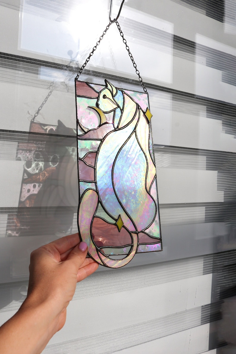 May include: A stained glass window hanging from a chain. The window features a colourful iridescent cat design with pink, blue and yellow accents.