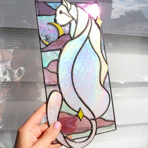 May include: A stained glass window panel depicting a white cat with iridescent glass, sitting against a background of pink, purple, and blue glass.