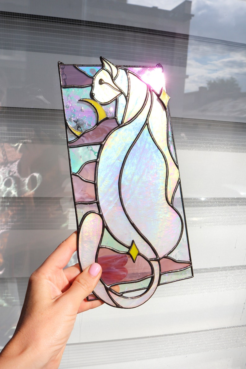 May include: A stained glass window panel depicting a white cat with iridescent glass, sitting against a background of pink, purple, and blue glass.