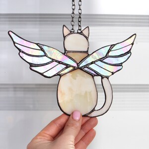 May include: A stained glass cat with iridescent wings hanging from a chain. The cat is white with a brown collar and is facing away from the viewer.