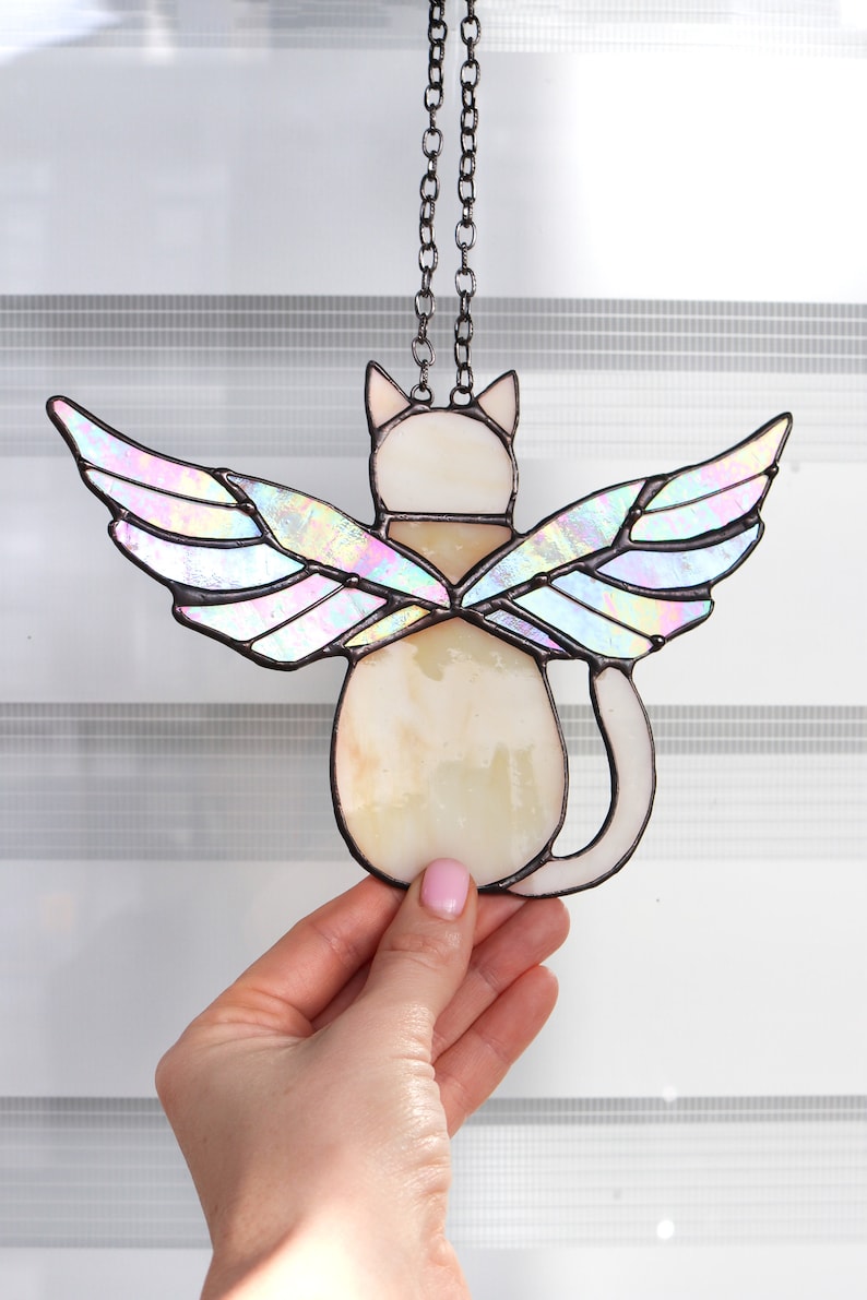 May include: A stained glass cat with iridescent wings hanging from a chain. The cat is white with a brown collar and is facing away from the viewer.