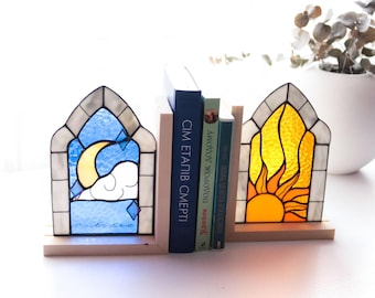Puede incluir: Soportes para libros de vidrieras con diseño de ventana. Uno presenta un cielo azul con luna y nubes, el otro un sol amarillo. Los sujetalibros son de vidrio y madera, sosteniendo varios libros.