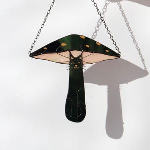 May include: A stained glass hanging decoration of a black cat sitting under a green mushroom with white spots. The mushroom has a pink underside.
