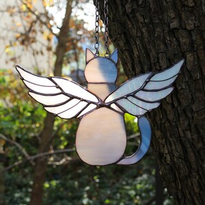 May include: A stained glass cat with wings hanging from a chain. The cat is white with a blue tail and iridescent wings.