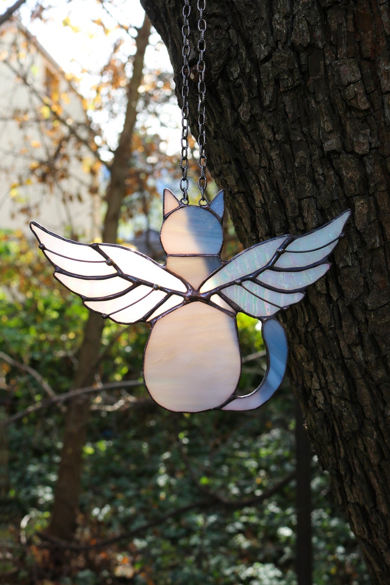 May include: A stained glass cat with wings hanging from a chain. The cat is white with a blue tail and iridescent wings.