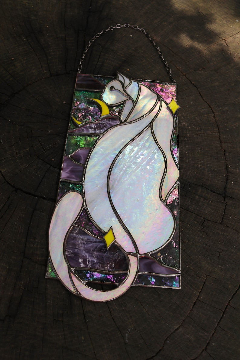 May include: A stained glass window hanging from a chain, featuring a white cat with iridescent glass, sitting with a crescent moon and stars in the background.