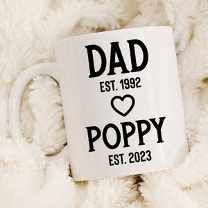 May include: White ceramic mug with black text that reads "DAD EST. 1992" with a heart symbol and "POPPY EST. 2023".