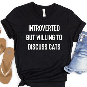 May include: Black t-shirt with white text that reads "Introverted but willing to discuss cats". The shirt is laid flat on a white background with a pair of brown flip flops and denim shorts.