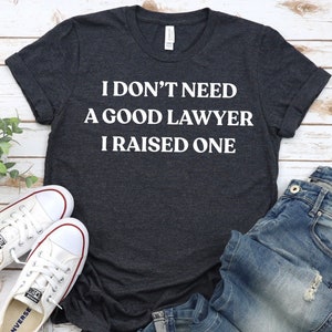 Puede incluir: Una camiseta gris oscuro con el texto "I don't need a good lawyer I raised one" en letras blancas. La camiseta está colocada sobre una superficie de madera blanca con un par de zapatos de lona blancos y unos vaqueros azules.