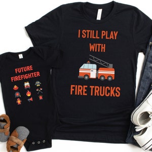 Puede incluir: Dos camisetas negras con temática de camión de bomberos. La camiseta más grande dice "Todavía juego con camiones de bomberos" y tiene un gráfico de camión de bomberos rojo. La camiseta más pequeña dice "Futuro bombero" y tiene una variedad de iconos relacionados con los camiones de bomberos.