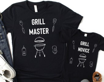 Könnte beinhalten: Zwei schwarze T-Shirts mit weißem Text und Illustrationen. Das größere Shirt sagt "Grill Master" und das kleinere Shirt sagt "Grill Novice". Beide Shirts zeigen einen Grill, einen Spatel, eine Zange und eine Flasche Barbecuesauce.
