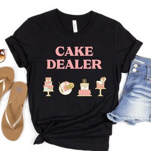 Puede incluir: Camiseta negra con el texto "CAKE DEALER" en letras rosas. La camiseta tiene cuatro peque&ntilde;as im&aacute;genes de pasteles en dorado y rosa.