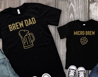 Könnte beinhalten: Zwei schwarze T-Shirts mit goldener Beschriftung. Das größere Shirt trägt die Aufschrift "Brew Dad" mit einem Bierkrug-Motiv. Das kleinere Shirt trägt die Aufschrift "Micro Brew" mit einem Hopfen-Motiv. Beide Shirts liegen auf einer Holzoberfläche mit Jeans und schwarzen Turnschuhen.