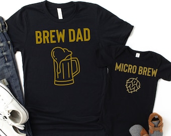 Könnte beinhalten: Zwei schwarze T-Shirts mit goldener Schrift und Grafiken. Das größere Shirt trägt den Schriftzug "Brew Dad" mit einer Bierkrüg-Grafik. Das kleinere Shirt trägt den Schriftzug "Micro Brew" mit einer Hopfen-Grafik.