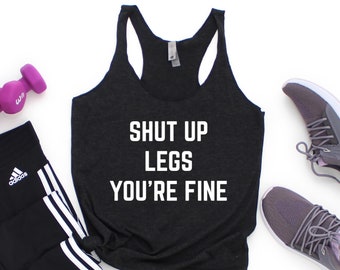 May include: A black tank top with the text "Shut Up Legs You're Fine" in white. The tank top is paired with black leggings with white stripes, purple dumbbells, and gray sneakers.